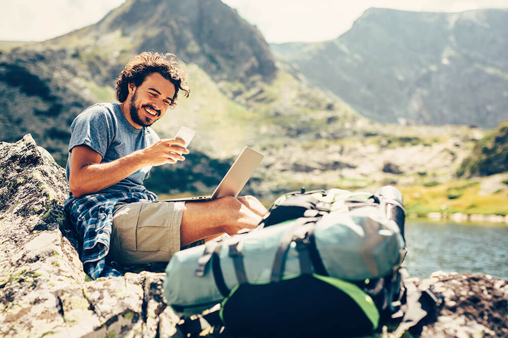 A man checks his laptop and phone while working remotely from a mountainous region