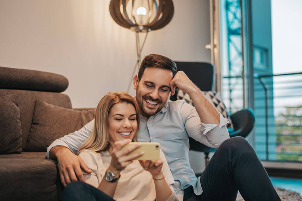 Couple sitting on the floor in front of the sofa laughing at the screen of the woman's phone.