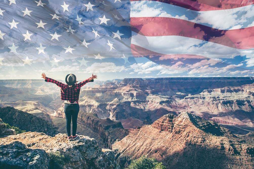 Woman stands on mountain with arms raised, an image of the American flag in the sky