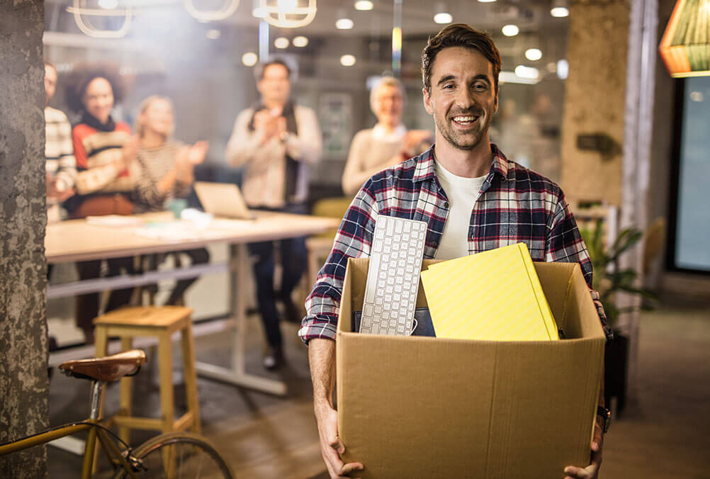 A smiling man carries a box of possessions out the door as he quits his job after winning the lottery