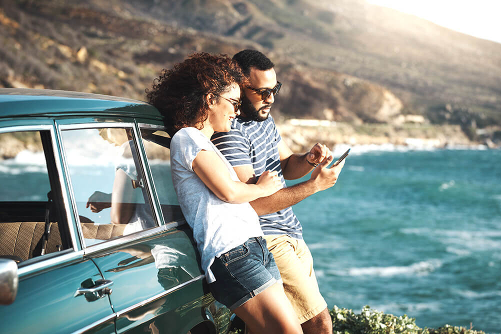 Couple checking the EuroMillions results on their phone next to the sea whilst leaning against a classic car