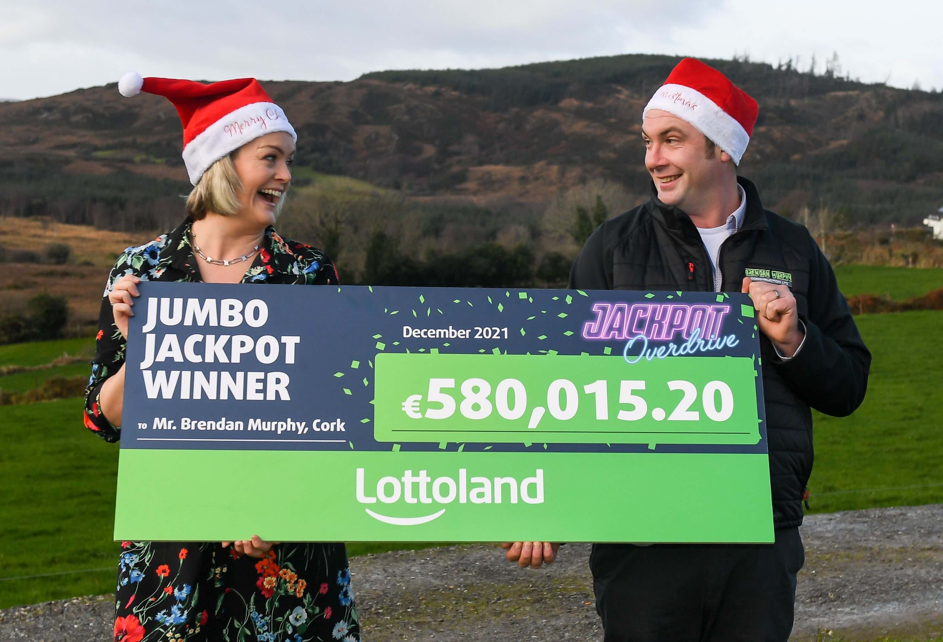 Lottoland winner holding up his cheque and smiling with his wife after winning the jackpot wearing Christmas hats to celebrate