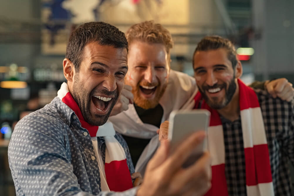 three guys with sports scarves betting on sports with lottoland online on their mobile